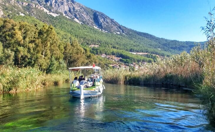 Akyaka’daki Azmak Nehri üzerinde ilerleyen tur teknesi; çevresi sazlıklarla çevrili, arkada yemyeşil orman ve dağ manzarası görülüyor.